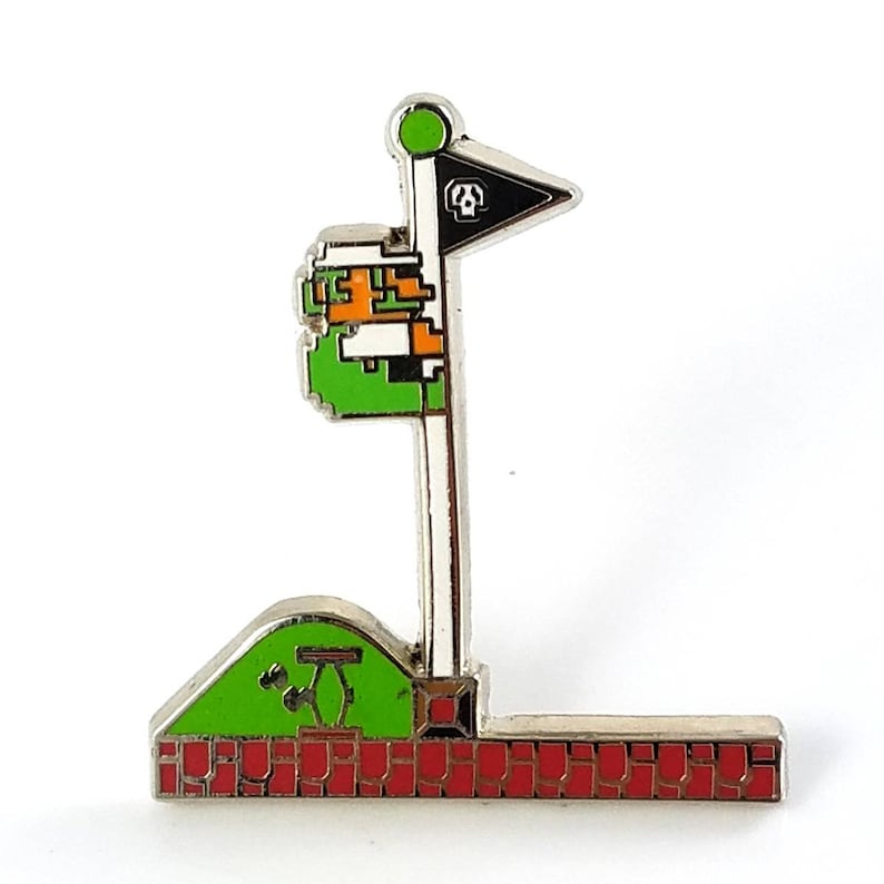 May include: A silver-colored pin featuring a pixelated golf flag design. The flag is black with a white skull and crossbones. The pin includes a green hill, red brick base, and a pixelated character. The flag pole has a green circle at the top.