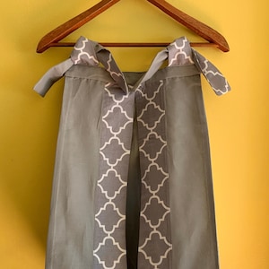 May include: A gray diaper caddy with white geometric pattern on the sides. The caddy has two ties at the top and a slit opening in the front.