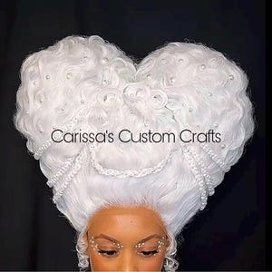 May include: A white wig styled in a heart shape, adorned with pearls and decorative elements. The text "Carissa's Custom Crafts" is displayed across the wig. The wig is worn by a person with decorative face jewels.