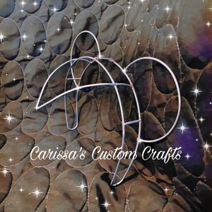 Pode incluir: Uma moldura de arame prateada em forma de lua crescente. A moldura está sobre um fundo de tecido acolchoado marrom. O texto "Carissa's Custom Crafts" é visível no centro inferior da imagem.