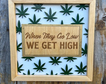 Pode incluir: Uma placa de madeira enquadrada com fundo branco e um padrão de folhas de maconha verdes. A placa tem uma placa de madeira com o texto "When They Go Low WE GET HIGH" em letras marrons.