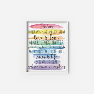 May include: A white framed print with a rainbow watercolor background and the text "I believe Immigrants make America great love is love Black Lives Matter women's rights are human rights All bodies are beautiful water is life Science is real and compassion is everything."