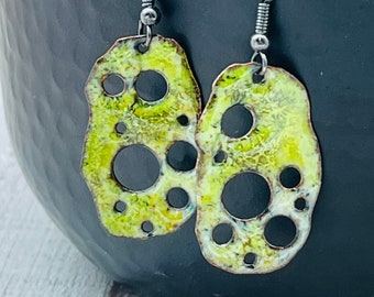 May include: A pair of green and white enamel earrings with a unique, organic shape and cut-out circles. The earrings are suspended from silver-toned hooks.
