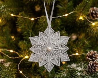 May include: A silver-colored, star-shaped snowflake ornament featuring intricate, lace-like embroidery patterns and a small, centered white pearl bead. The ornament hangs from a thin, matching silver ribbon loop against a green pine tree background.