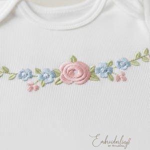 Può includere: Body da bambino bianco con ricamo floreale. Il ricamo presenta una rosa rosa, fiori blu e foglie verdi. Il body ha una trama a coste e uno scollo tondo. Il marchio "Embroiderlings" è scritto in corsivo in basso.