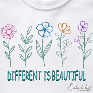 以下が含まれることがあります： 白い生地に刺繍された花の模様。デザインは、緑の茎と葉を持つ5つのカラフルな花が特徴です。「DIFFERENT IS BEAUTIFUL」というテキストが、ターコイズ色で花の下に刺繍されています。「Embroiderdarling」のブランド名が右下にあります。