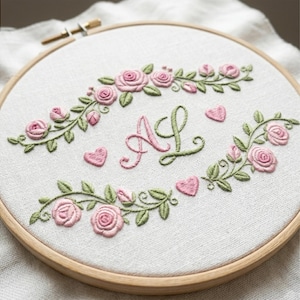 Può includere: Un telaio da ricamo circolare in legno contenente tessuto bianco con rose rosa, foglie verdi e piccoli cuori rosa ricamati a mano. Il centro presenta le lettere AL ricamate in corsivo con dettagli in filo rosa e verde.