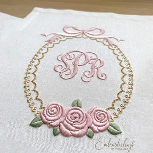 Könnte beinhalten: Gesticktes Design auf weißem Stoff mit einem rosa Blumenkranz mit den Initialen "SR". Der Kranz enthält eine Schleife, Rosen und grüne Blätter. Der Text "Embroiderlings" befindet sich unten.