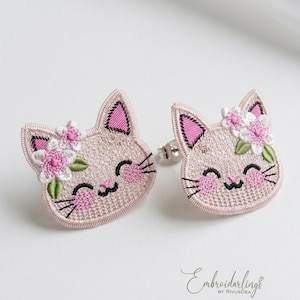 Pode incluir: Um par de brincos de tarracha com rostos de gato bordados em fio rosa claro. Cada gato tem o interior das orelhas rosa, bochechas rosadas, bigodes pretos e um sorriso. Um grupo de flores brancas e rosas com folhas verdes adorna uma orelha.