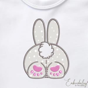 Pode incluir: Desenho bordado da parte traseira de um coelho cinza com pés cor-de-rosa e uma cauda branca e fofa. O coelho está sobre um fundo branco.
