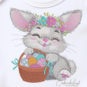 May include: Embroidered design of a smiling bunny wearing a floral crown and holding a basket of Easter eggs. The bunny is gray with pink accents on the ears and nose. The basket is brown with blue ribbons.