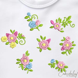 May include: A white fabric with embroidered floral designs in pink, yellow, blue, and green. The flowers are arranged in various clusters and patterns.