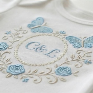 May include: White onesie with light blue embroidered butterflies, roses, and foliage. The design includes a circular frame with the embroidered letters "C&L". The embroidery is detailed and delicate, creating a soft, elegant look.
