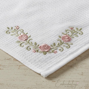 May include: White hand towel with an embroidered floral design. The embroidery features pink roses, green leaves, and delicate swirls. The towel has a textured surface and a folded corner.