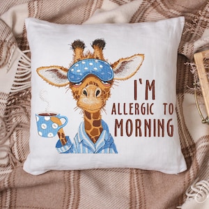 Puede incluir: Almohada blanca con una jirafa bordada que lleva una máscara para dormir azul y pijama. La jirafa sostiene una taza de café azul y blanca. El texto "Soy alérgico a la mañana" está bordado en la almohada.
