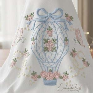Puede incluir: Tela blanca bordada con un diseño de globo aerostático azul claro, con rosas rosas y un lazo. Mariposas rosas y detalles florales rodean el globo. El diseño incluye el texto "Embroiderlings".