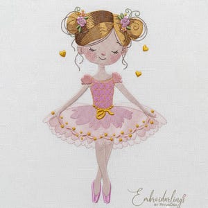 May include: Embroidered image of a ballerina in a pink tutu and pointe shoes. The ballerina has her hair in two buns with floral accents. Gold hearts float around her. The text "Embroiderlings" is at the bottom.