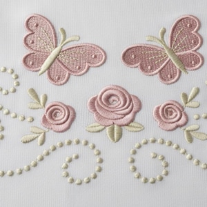 Può includere: Design ricamato con due farfalle rosa e tre rose rosa con foglie verde chiaro. Una fila di piccole perline bianche sporche si curva lungo il fondo. Il design è su sfondo bianco.