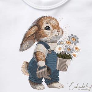 Op de afbeelding: Borduurwerk van een bruin konijn dat een blauwe overall draagt en een gieter en een boeket witte bloemen vasthoudt.