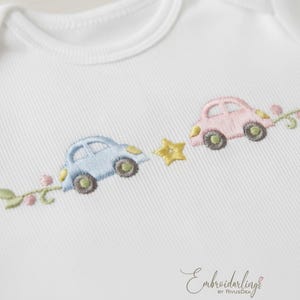 Puede incluir: Body de bebé blanco con detalles bordados. Presenta un coche azul claro, un coche rosa, una estrella amarilla y detalles florales. El diseño está bordado en colores pastel, creando una estética suave. El texto "Embroiderlings" está en la parte inferior.