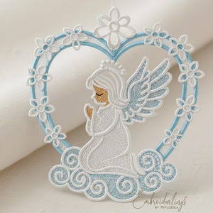 Puede incluir: Adorno de ángel bordado en blanco y azul claro. El ángel está arrodillado sobre una nube dentro de un marco en forma de corazón adornado con flores. El diseño presenta bordados detallados y una estética delicada.