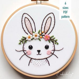 May include: Embroidered bunny rabbit with a floral crown in a wooden embroidery hoop. The bunny has pink cheeks and black eyes. The floral crown is made of white and pink flowers with green leaves. The text "Embroidering by RivusDea" is in the bottom right corner of the image.