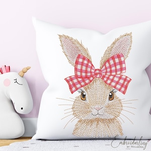 Puede incluir: Cojín cuadrado blanco con un bordado de la cara de un conejito con un lazo de cuadros rojos y blancos. El conejito tiene ojos marrones y bigotes. Un artículo decorativo, posiblemente para un dormitorio infantil.