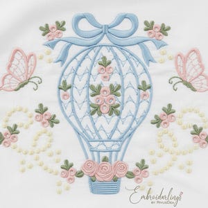 May include: Embroidered design featuring a light blue hot air balloon with pink roses and green leaves. Two pink butterflies and floral accents surround the balloon. The design includes a light blue bow at the top and a pearl-like border.