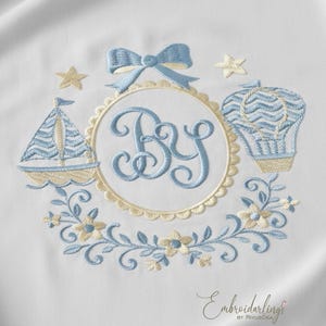 Può includere: Design ricamato con una barca a vela, mongolfiera, stelle ed elementi floreali in azzurro e beige. Un monogramma centrale con le lettere "BY" è circondato da un cerchio smerlato e un fiocco blu.