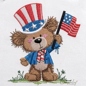 Op de afbeelding: Geborduurde teddybeer met een patriottische hoge hoed en blauwe jas, met een kleine Amerikaanse vlag. De beer heeft een rood-wit-blauwe vlinderdas en staat op een stukje groen gras. Een charmant ontwerp voor patriottische vieringen.