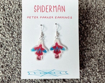 Puede incluir: Un par de pendientes de plata con imágenes de Spiderman en rojo, azul y blanco. La tarjeta dice "Spiderman Peter Parker Earrings".