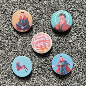 May include: Five round buttons with different designs. One button features a cartoon character in a red and blue suit with the text "Sanctum Santorum New York Hong Kong London". Another button features a cartoon character in a red and blue suit with a yellow background. The remaining three buttons feature a cartoon character in a red and blue suit with a blue background.
