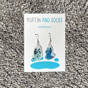 May include: A pair of earrings featuring cartoon dogs, one blue and one black and white, with the text "MUFFIN AND SOCKS EARRINGS" on a white card.