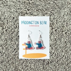 May include: A pair of Paddington Bear earrings. The earrings are made of clear acrylic and feature a cartoon image of Paddington Bear wearing a blue coat and red boots. The earrings are hanging from silver hooks.