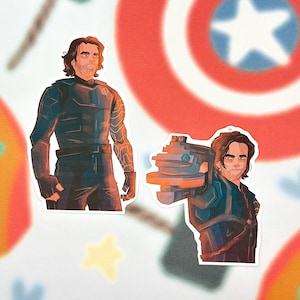 May include: Two stickers featuring a character in a dark blue outfit. One sticker shows the character standing, and the other shows the character holding a weapon. The background has a red, white, and blue theme with star and shield designs.