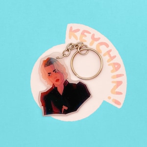 Op de afbeelding: Sleutelhanger met een illustratie van een persoon met blond haar en rode lippen, gekleed in een zwart topje. De sleutelhanger heeft een goudkleurige ring en ketting. Het woord "KEYCHAIN!" staat in een tekstballon.