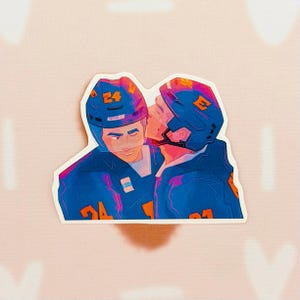May include: A sticker featuring two hockey players in blue uniforms and helmets, one kissing the other on the cheek. The sticker has a white border and is set against a pink background.