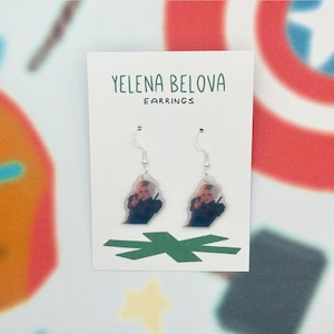 May include: Clear acrylic earrings featuring a character from the movie "Black Widow" holding a gun. The earrings are on a white card with the text "Yelena Belova Earrings". The earrings hang from silver-colored hooks.