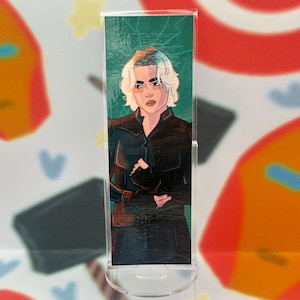 May include: A clear acrylic bookmark featuring a stylized illustration of a person with short, wavy, light blonde hair. The person is wearing a black outfit. The background is a textured green pattern.