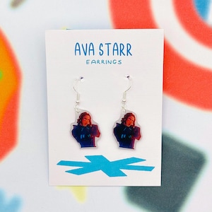 Può includere: Un paio di orecchini con una grafica di una persona con i capelli rossi e un abito blu. Gli orecchini sono su una carta bianca con il testo "AVA STARR EARRINGS" in blu. Lo sfondo ha un design astratto colorato.