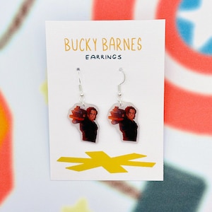 May include: A pair of dangle earrings featuring a graphic of a person carrying a stack of items. The earrings are on a white card with the text "BUCKY BARNES EARRINGS" in orange. A yellow arrow design is at the bottom of the card.