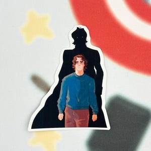 Könnte beinhalten: Ein Aufkleber mit einer Illustration einer Person in einem blauen Pullover und braunen Hosen, mit einer schwarzen Silhouette dahinter. Der Hintergrund umfasst ein rot-weißes Design und einen gelben Stern.