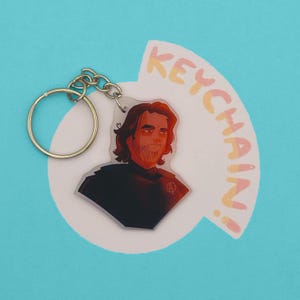 Op de afbeelding: Een heldere acryl sleutelhanger met een zilveren ketting en sleutelring. De sleutelhanger heeft een illustratie van een persoon met lang haar en een baard. Het woord "KEYCHAIN!" is geschreven in oranje en geel.