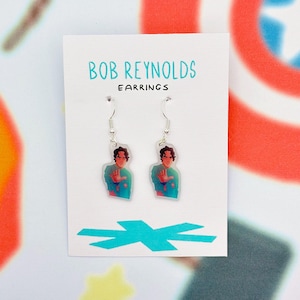 May include: A pair of dangle earrings featuring a cartoon image of a person in a teal shirt. The earrings are on a white card with the text "BOB REYNOLDS EARRINGS" in teal. The background has a colorful abstract design.