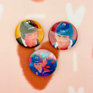 May include: Three round hockey-themed buttons. Each button features a stylized illustration of a hockey player wearing a helmet. The buttons are in shades of yellow, red, and blue. The player's jersey number is visible on two of the buttons.