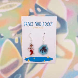 May include: A pair of dangle earrings on a white card with the text "GRACE AND ROCKY EARRINGS". One earring features a cartoon figure in red pants, the other a dark insect in a blue hexagon. Silver hooks.