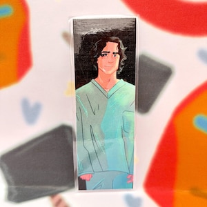 May include: A bookmark featuring an illustration of a person with dark curly hair wearing a light teal v-neck shirt and pants. The bookmark is in a clear plastic sleeve, with a colorful background.