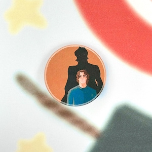 Könnte beinhalten: Ein runder Button mit einer Cartoon-Illustration einer Person in einem blauen Hemd mit einer dunklen Silhouette dahinter. Der Button hat einen orangefarbenen Hintergrund und befindet sich vor einem weiß-roten Hintergrund.