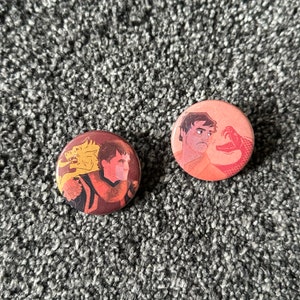 以下が含まれることがあります： イラストが描かれた2つの丸いピンバッジ。1つは金のドラゴンを持つ男性、もう1つは赤いヘビに面した男性が描かれています。ピンバッジは、グレーのテクスチャ加工された表面にあります。