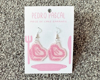 May include: A pair of pink acrylic earrings shaped like a piece of cake with the text "Pedro Pascal" on them. The earrings are on a white card with the text "Pedro Pascal Piece of Cake Earrings".